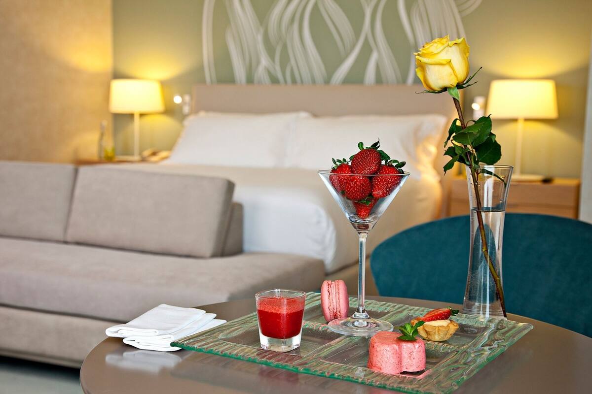 A hotel room with a bed, a bedside table with a glass of red drink, a glass of water, a vase with a yellow rose, and a plate with pastries and strawberries.