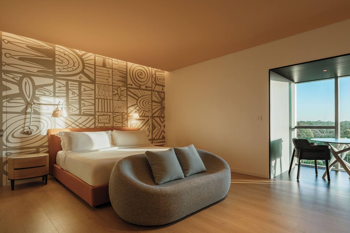 A modern bedroom with a large bed, a unique wall art design, a cozy seating area, and a balcony with a view.