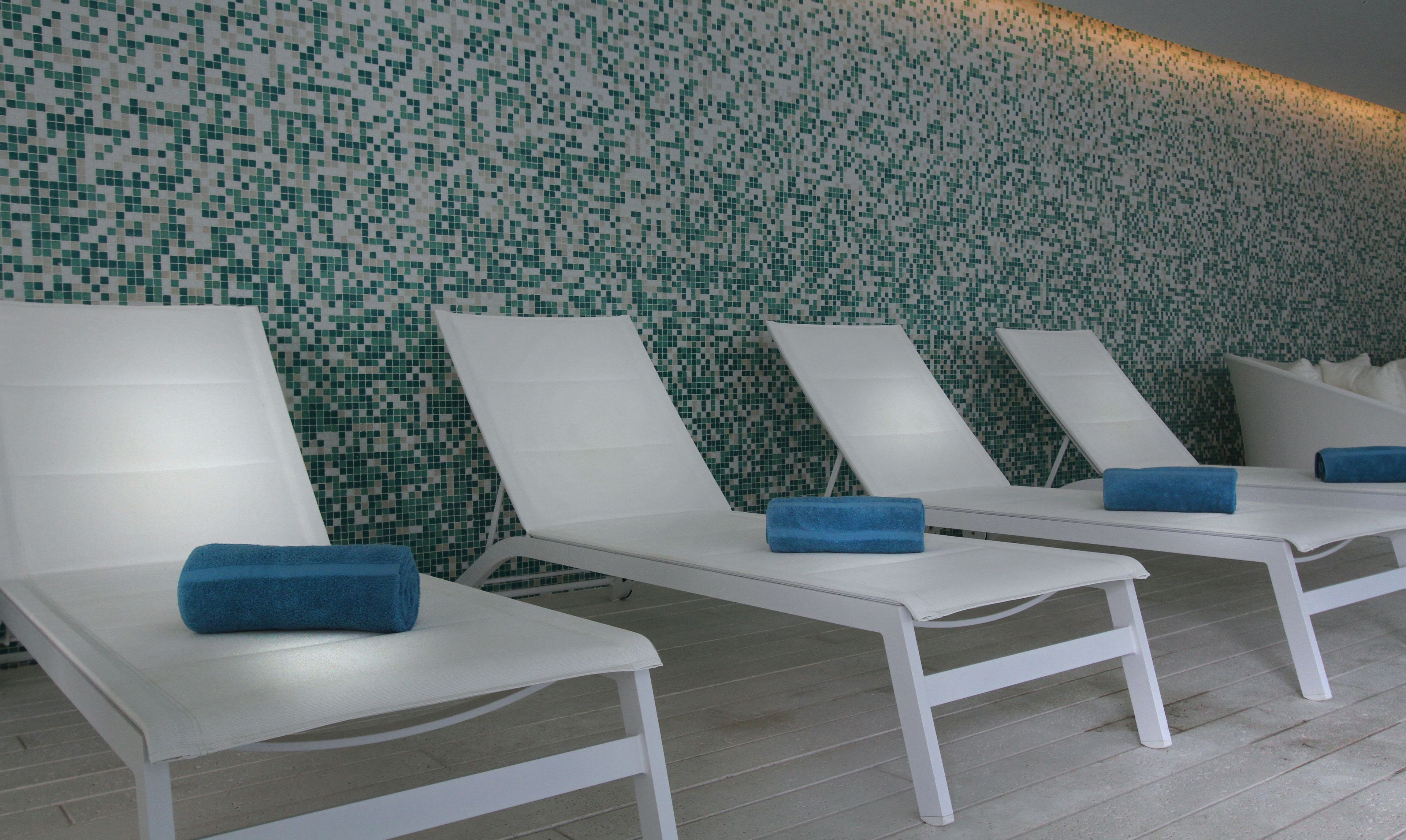 A row of white lounge chairs with blue towels, set against a wall with a green mosaic pattern.