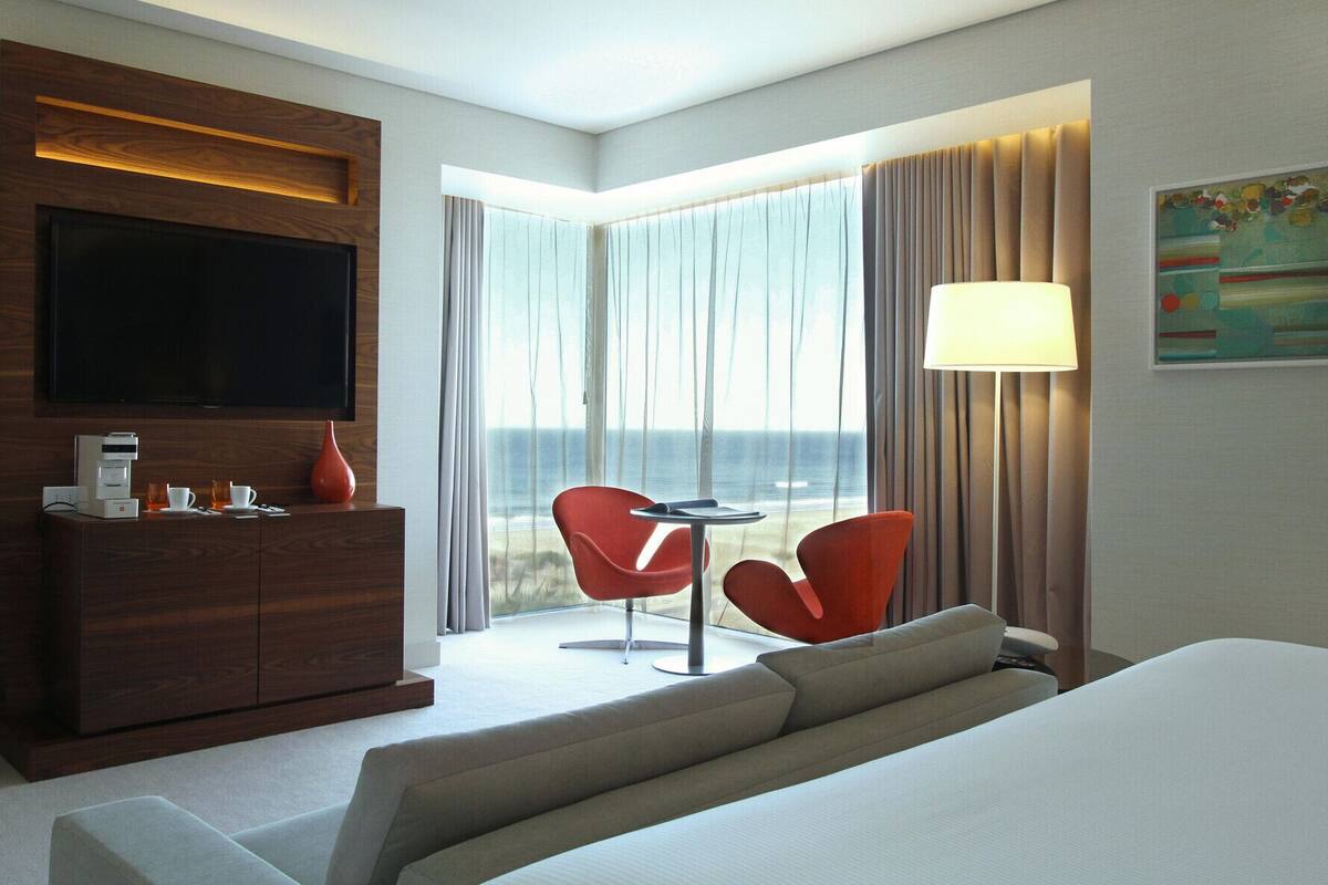 A hotel room with a bed, a TV, a coffee maker, a table with two chairs, and a view of the ocean.
