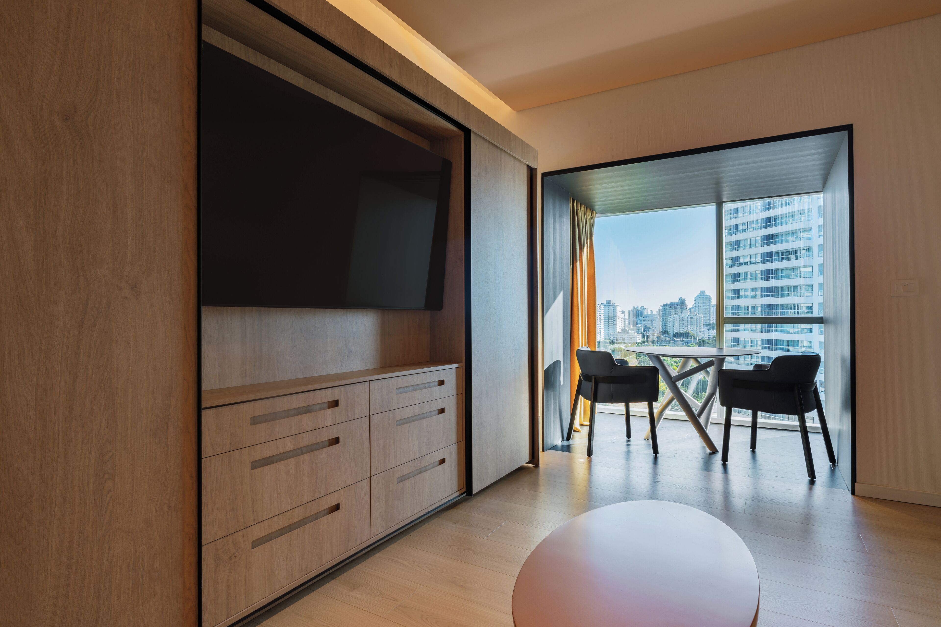 A modern living room with a flat-screen TV, wooden cabinets, a round table, and a view of a cityscape through large windows.