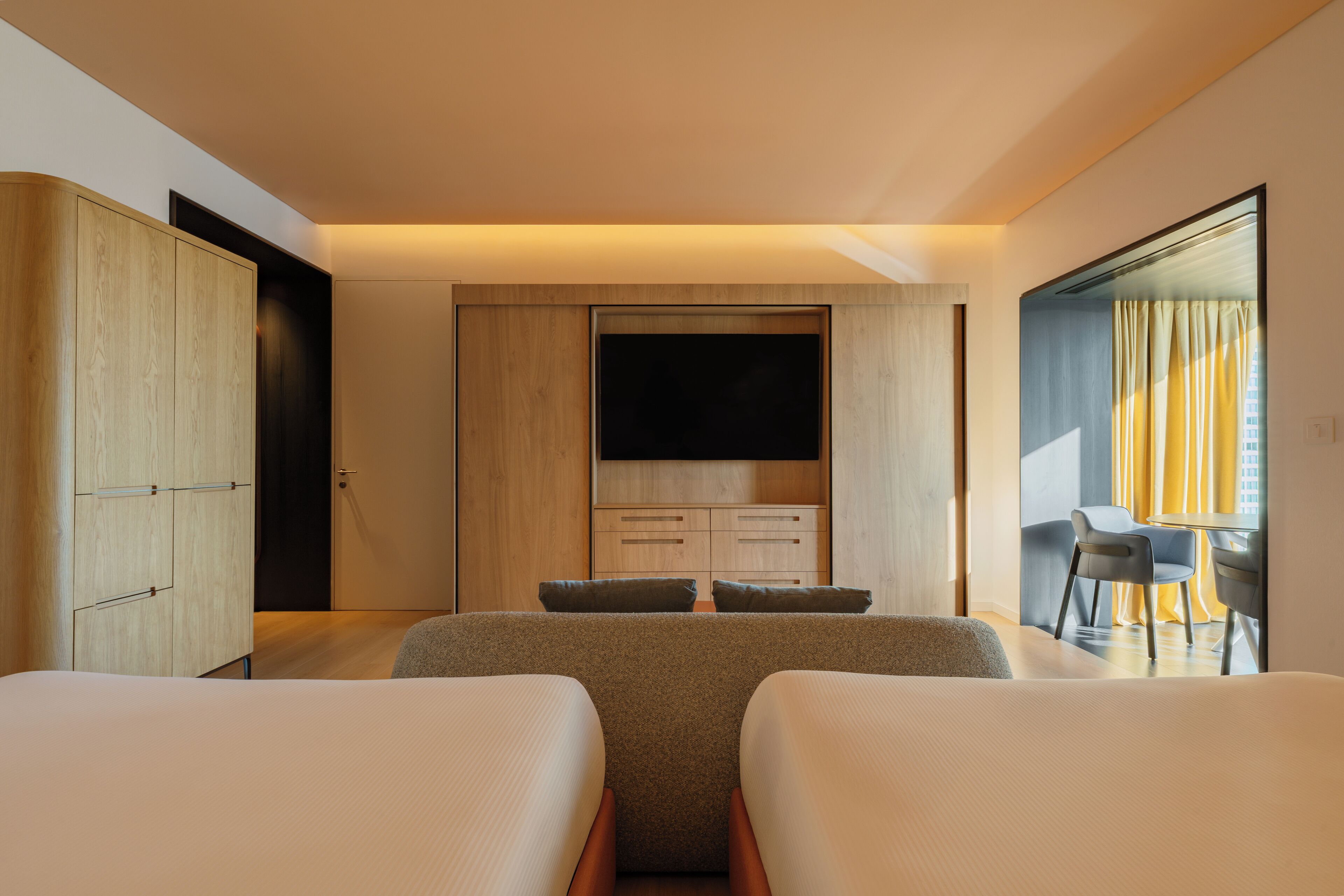 A modern hotel room with two beds, a TV, a desk, and a view of the city.