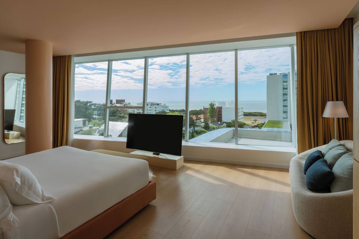 A hotel room with a large bed, a TV on a stand, a sofa, and a large window offering a city view.
