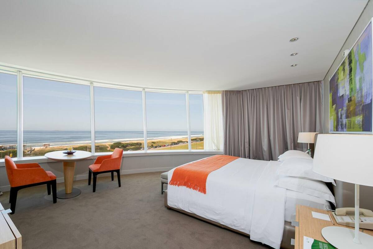 A hotel room with a large window, a bed, a desk with a lamp, and two chairs.