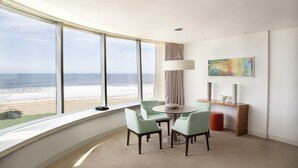 A modern hotel room with a large window overlooking the beach, a round table with chairs, a lamp, and a painting on the wall. - The Grand Hotel Punta Del Este (Punta del Este)