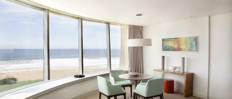Una habitación de hotel moderna con un amplio ventanal con vista a la playa, una mesa redonda con sillas, una lámpara y un cuadro en la pared.