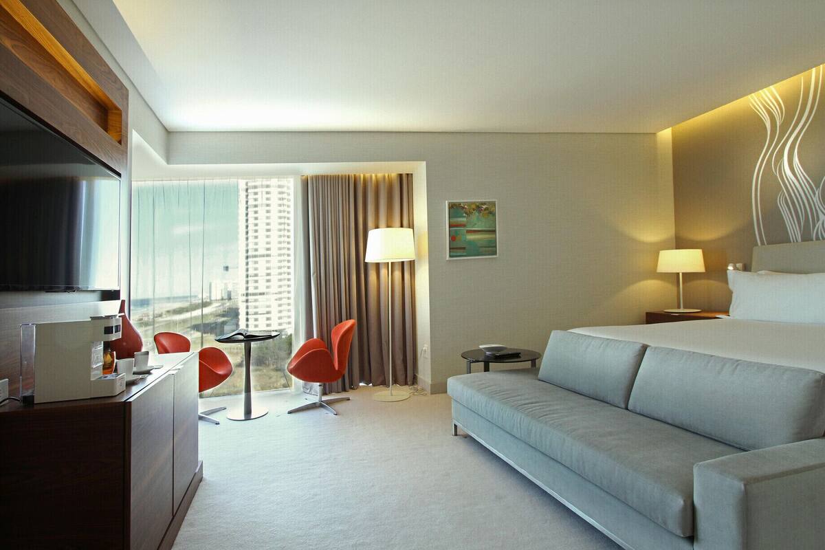 A hotel room with a bed, a desk with a TV, a sofa, a chair, a small table, and a window with curtains.
