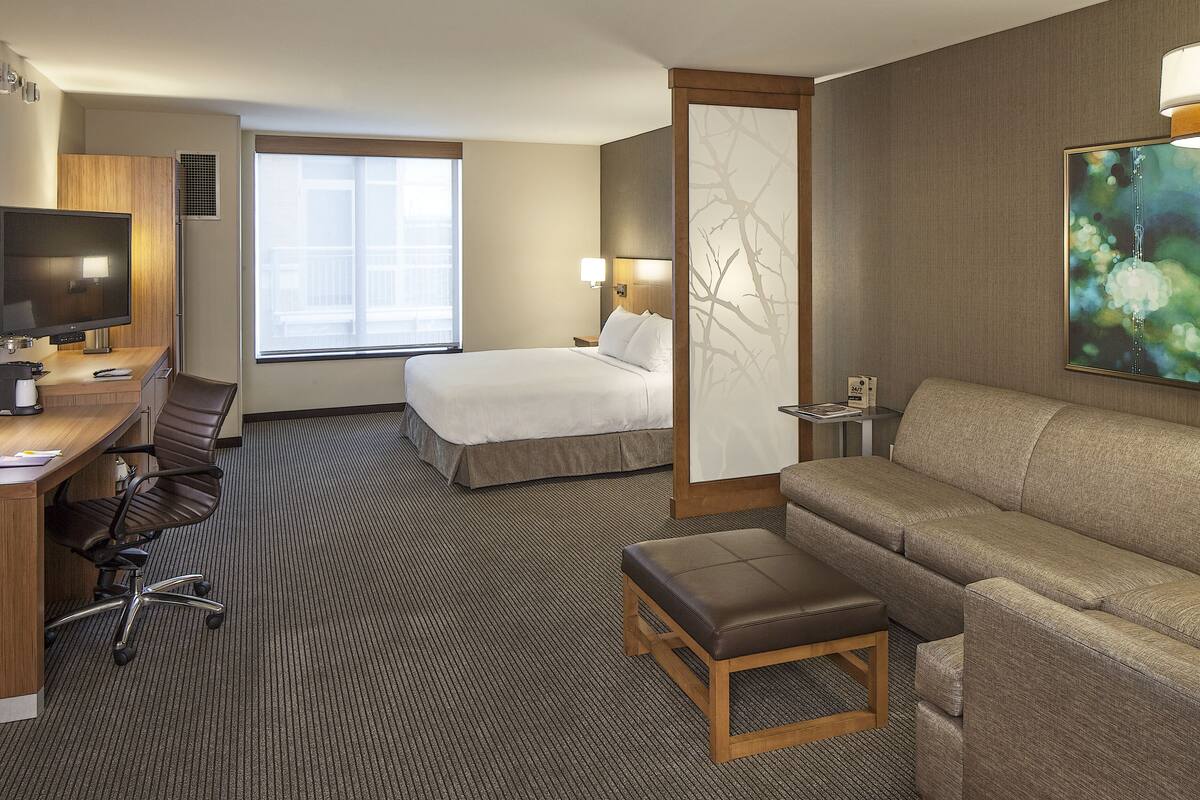 Une chambre d’hôtel avec un lit, un bureau équipé d’un ordinateur, une chaise, un canapé et une grande fenêtre.