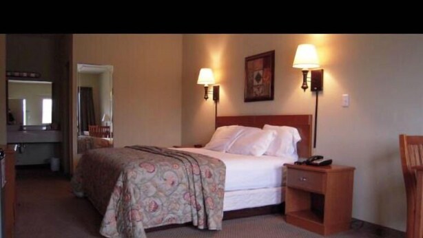 A hotel room with a bed, bedside tables, a nightstand, a mirror, and a door leading to another room. - City Heart Inn & Suites (Piedmont)