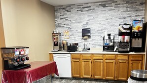 A compact kitchen with a stone backsplash, wooden cabinets, a coffee station, and a table with a red tablecloth. - Designer Inn & Suites (Toledo)