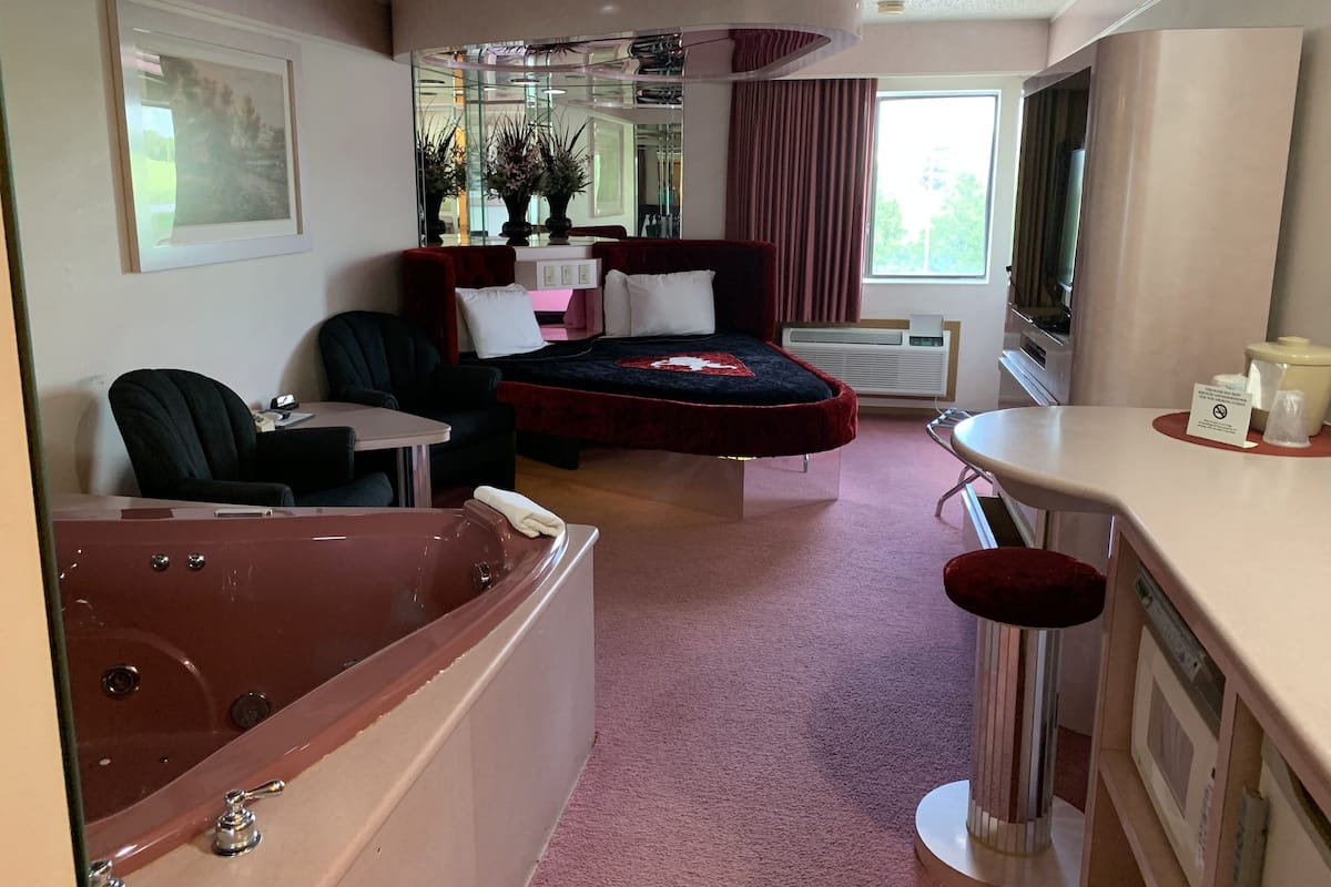 Quarto de hotel compacto com uma cama, uma pequena mesa de escritório, uma cadeira, uma banheira e uma janela com cortinas.