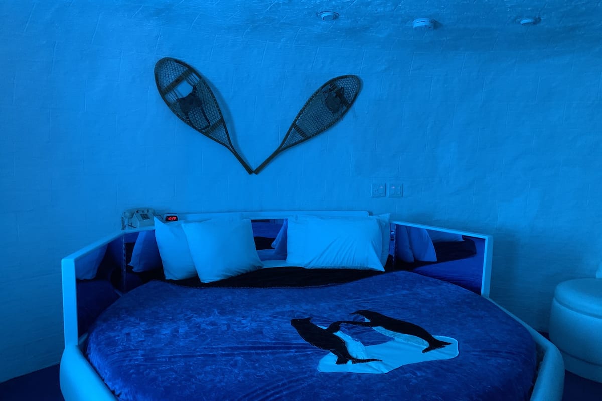 Quarto com cama, almofadas, decoração em formato de coração na parede e mesa de cabeceira.