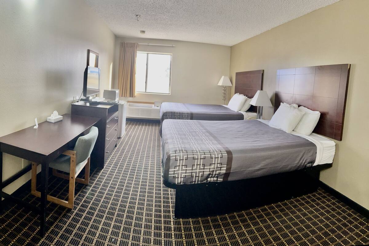 Quarto de hotel com duas camas, uma mesa, uma cadeira, uma televisão e uma janela com cortinas.