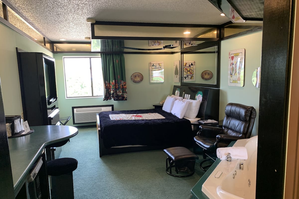Quarto de hotel com cama, escrivaninha, cadeira, banco, banheira e janela com cortinas.