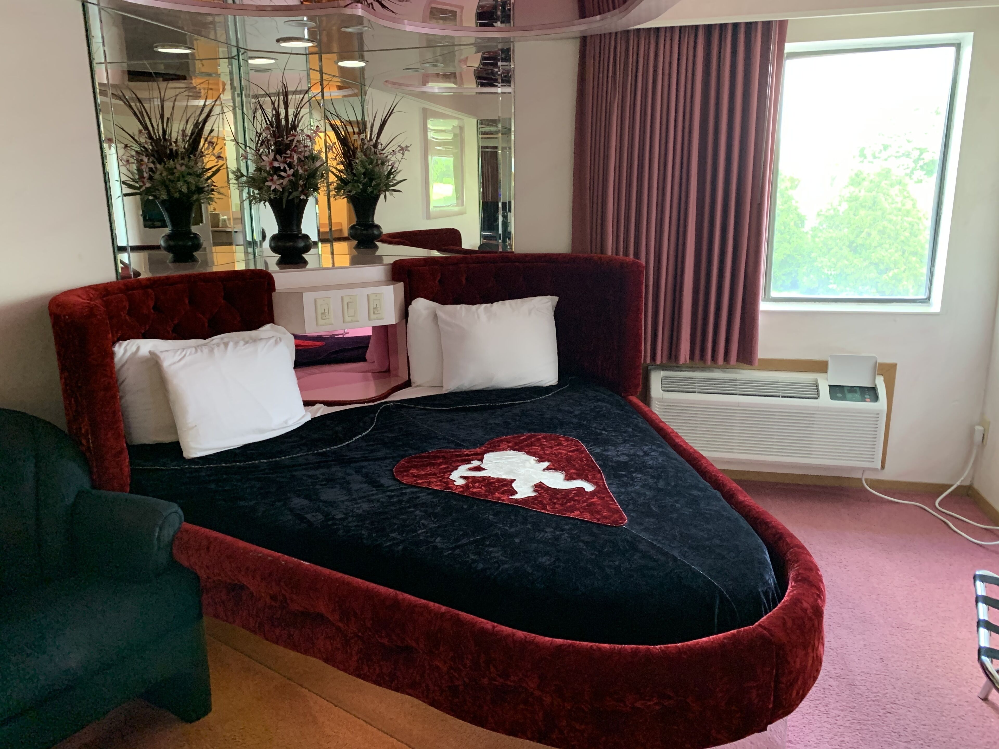 A modern bedroom with a large bed, a red upholstered headboard, a bedside table with lamps, and a window with curtains.