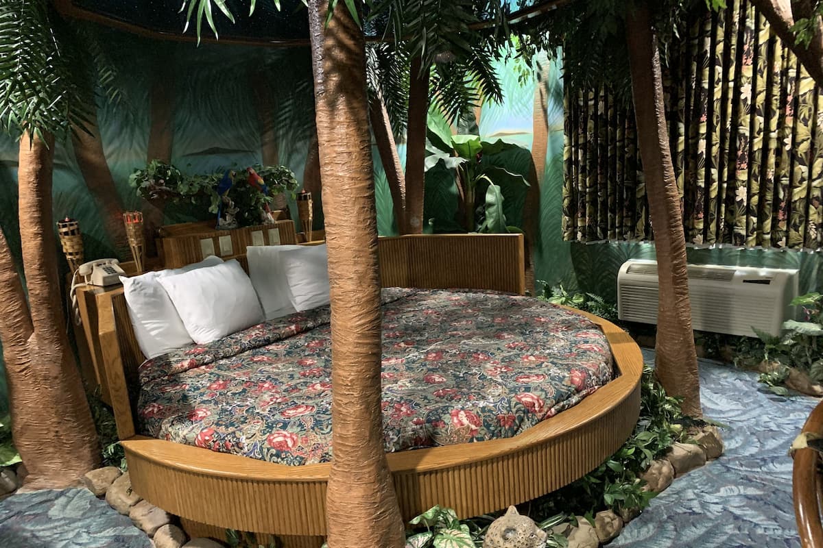 Uma cama redonda com colcha florida, cercada por um estrutura de madeira e plantas tropicais.