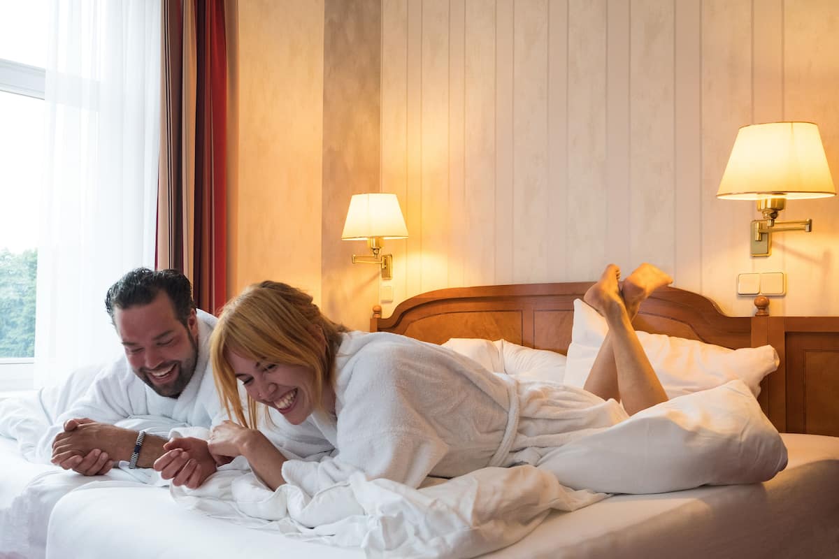 Ein Paar in weißen Bademänteln liegt auf einem Bett mit einem hölzernen Kopfstück, zwei Nachttischlampen und einem Fenster mit Vorhängen.