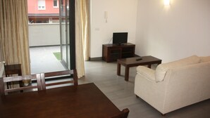 Una sala de estar con un sofá, un televisor sobre un mueble bajo, una mesa de comedor con sillas y vistas a un edificio a través de una puerta de vidrio.