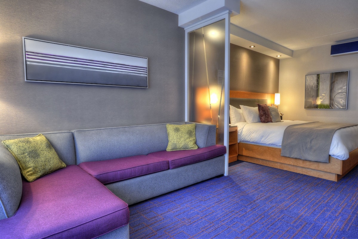 A modern hotel room with a gray sofa, a bed, and a large window.