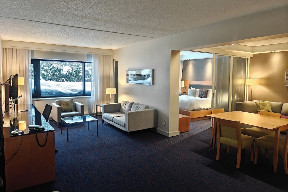 A hotel room with a large window, a sofa, a dining table, and a bed.