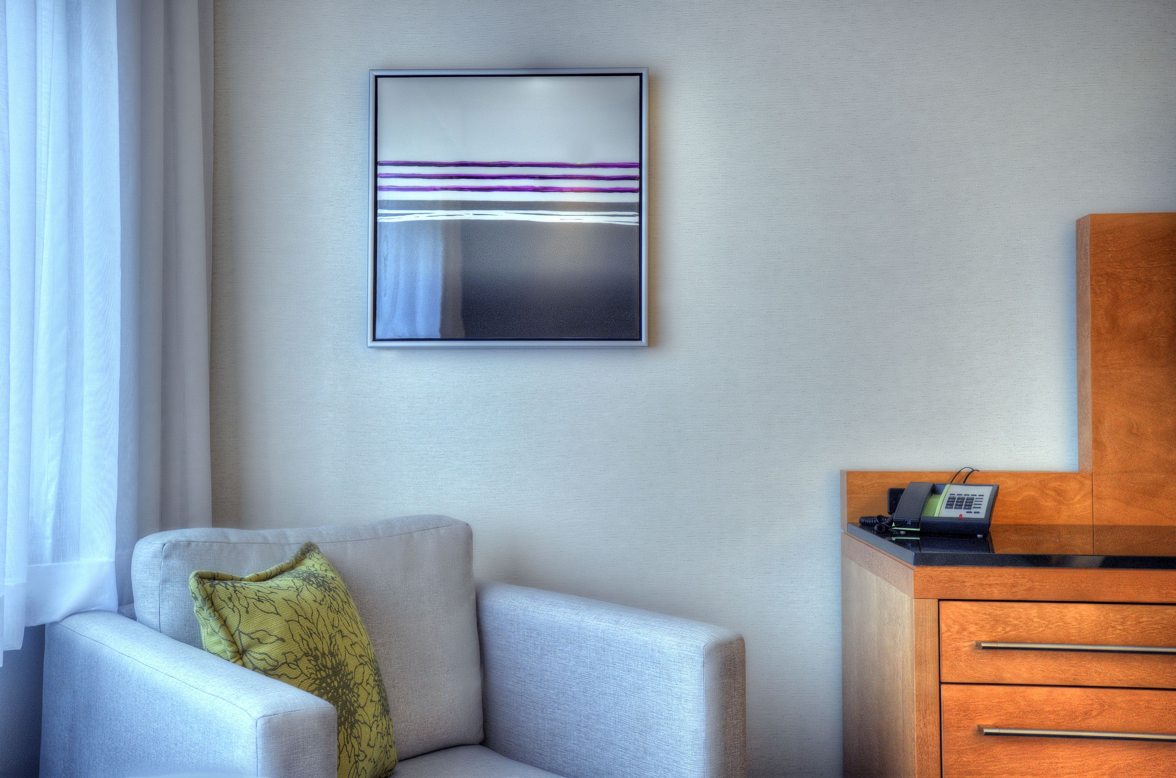 A hotel room with a grey armchair, a wooden dresser, and a framed abstract artwork on the wall.