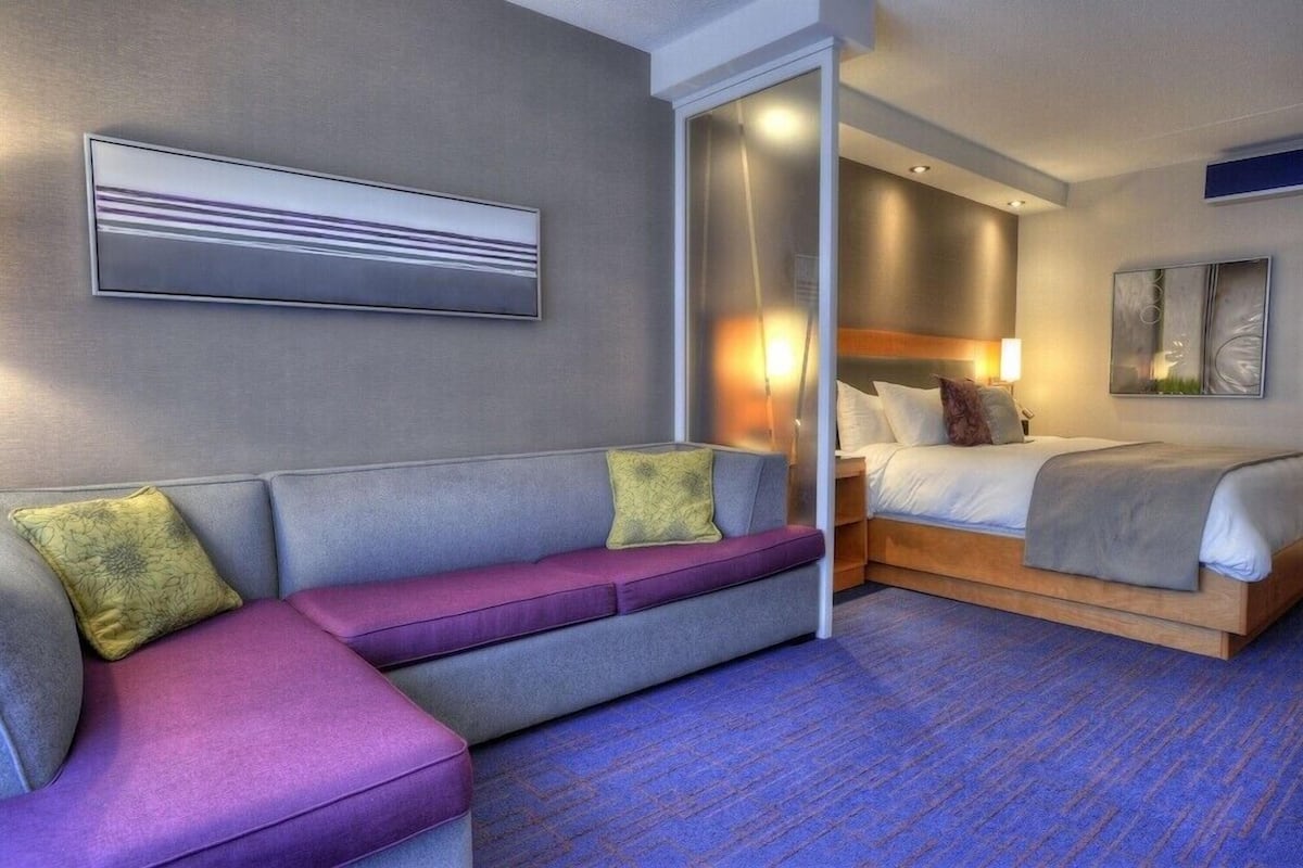 A modern hotel room with a grey sofa, a bed with a wooden frame, and a blue carpet.