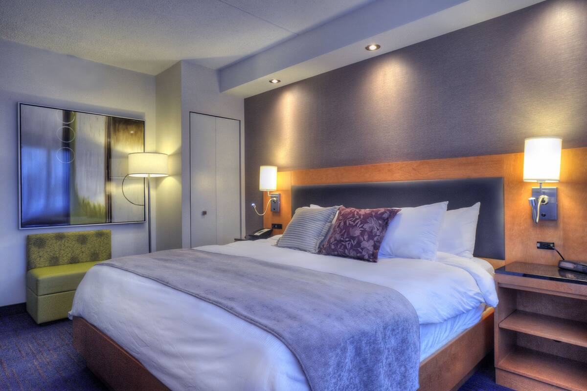 A hotel room with a large bed, a bedside table, a lamp, a mirror, and a small seating area.