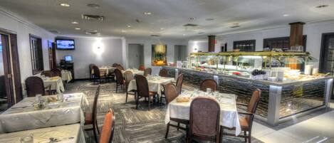 A hotel dining area with multiple tables, chairs, a buffet counter, and a television screen.