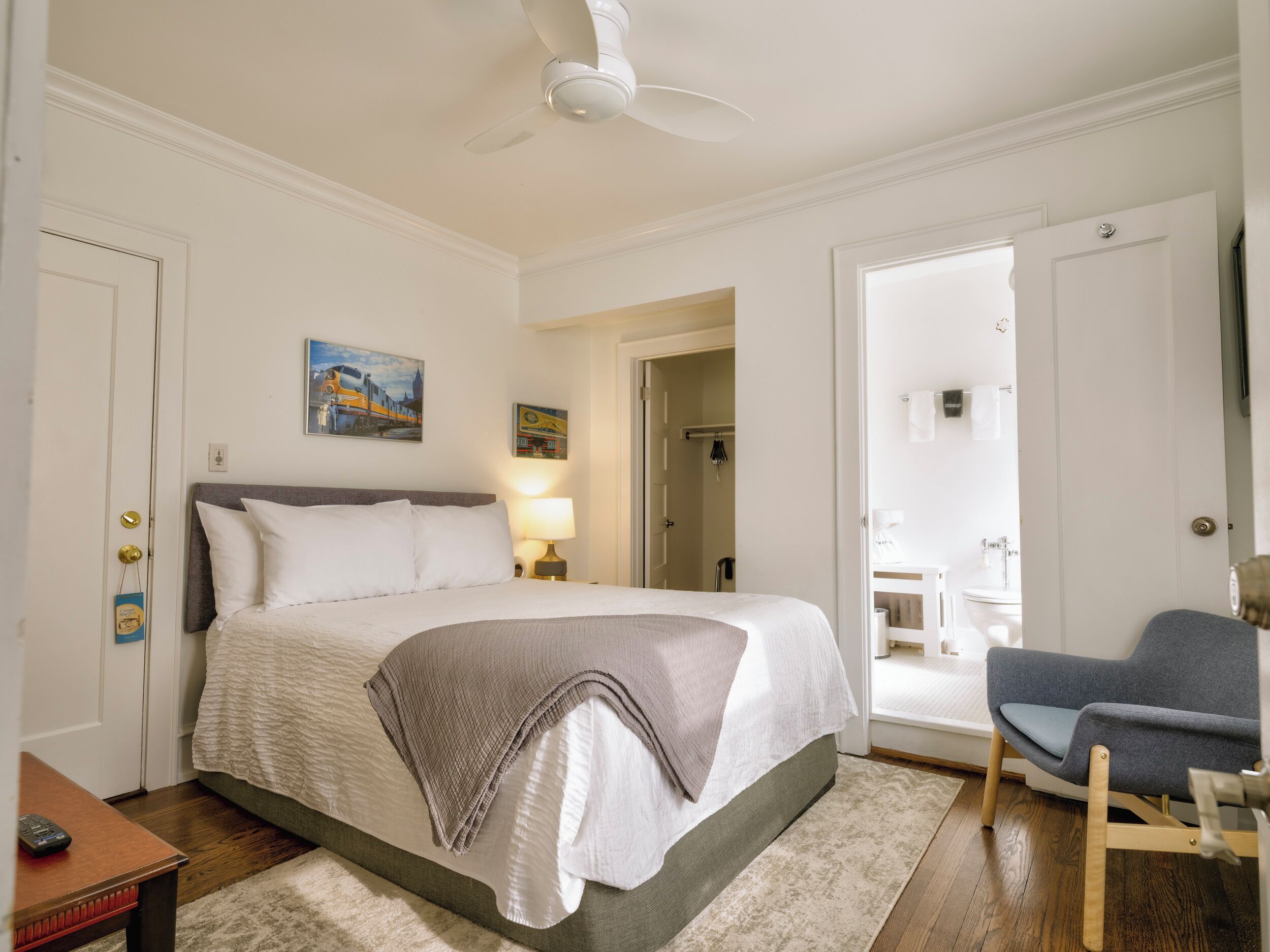 Habitación de hotel con cama, escritorio, silla, ventilador de techo y baño visible a través de una puerta abierta.