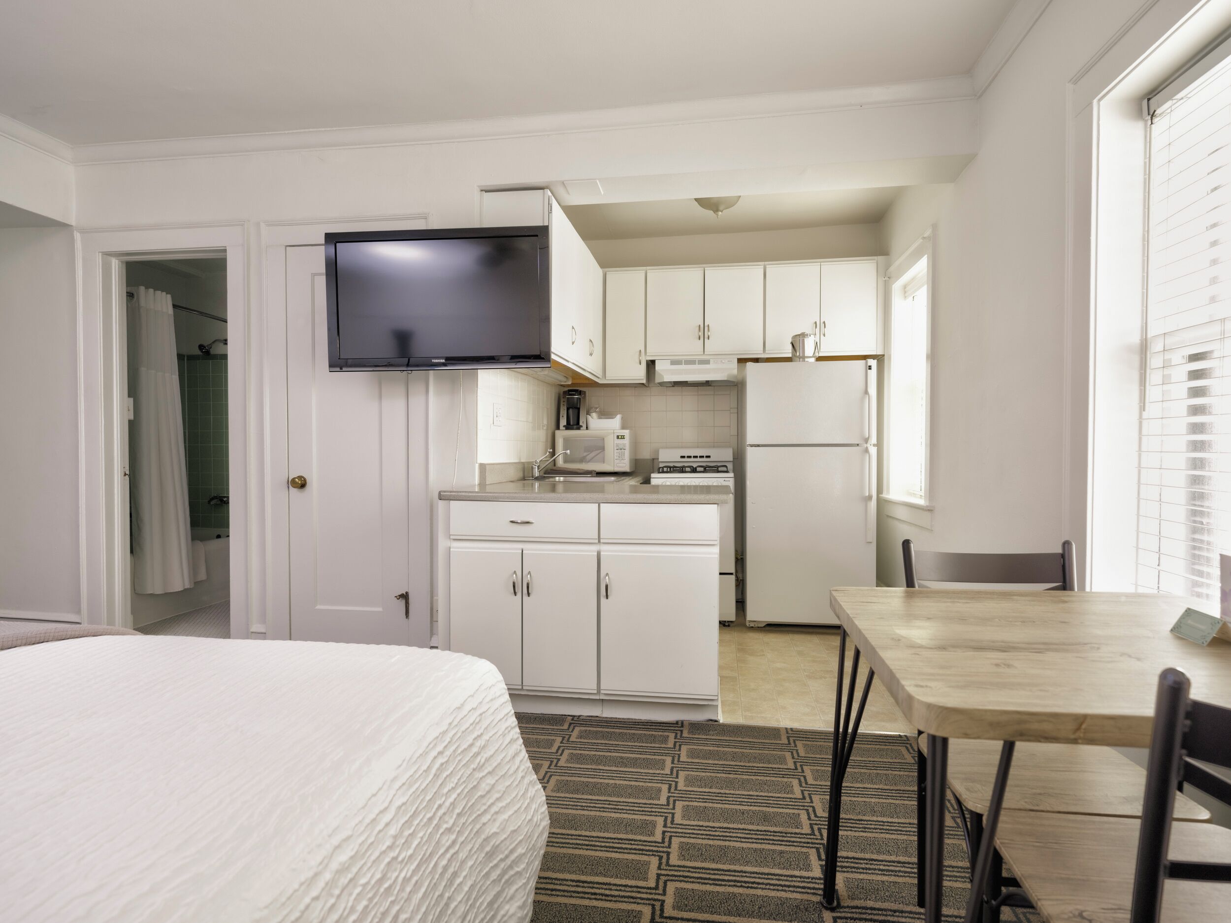 Une chambre d’hôtel compacte comprenant un lit, une kitchenette, une table à manger et un téléviseur fixé au-dessus du comptoir.