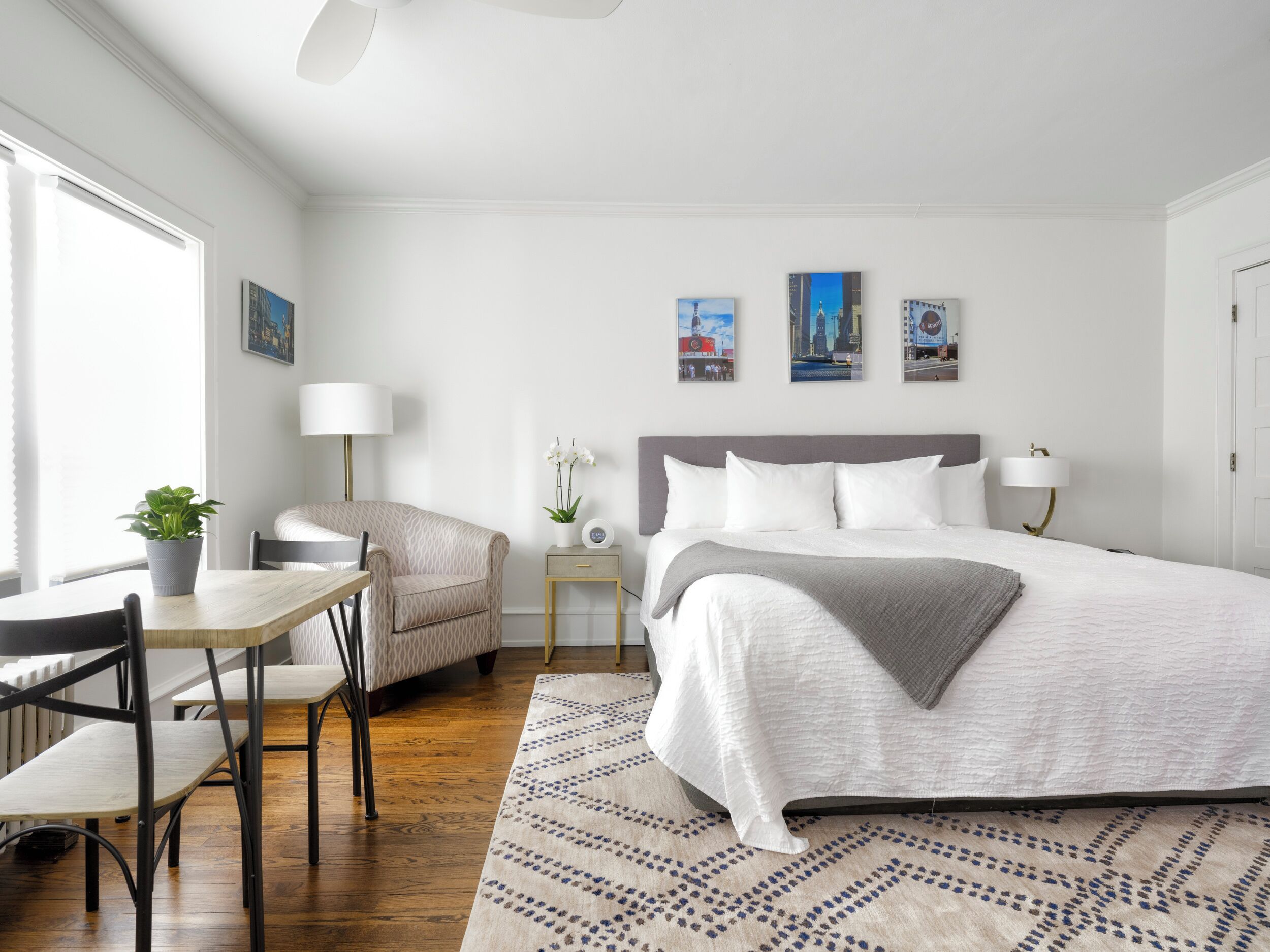 A modern hotel room with a large bed, a dining area with a table and chairs, a small side table with a lamp, and artwork on the walls.