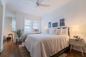 Un dormitorio con cama, mesita de noche, ventilador de techo, planta en maceta, ventana con persianas y una obra de arte en la pared.