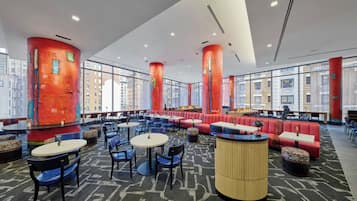 Un vestíbul d'hotel modern amb taules rodones, seients vermells i grans finestres que donen vistes a la ciutat.