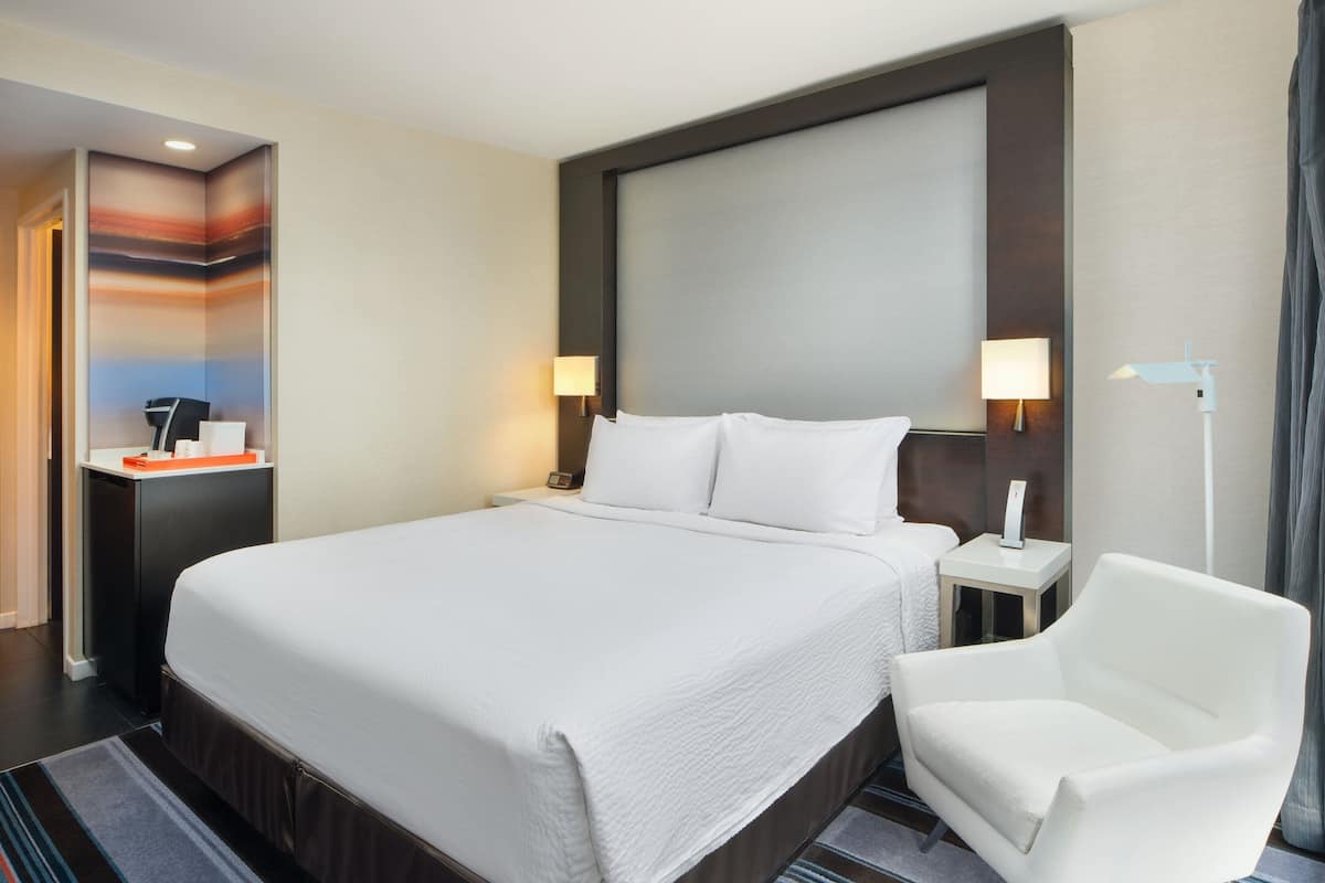 Habitación de hotel con una cama grande, una mesita de noche con lámpara, una silla y vistas a la ciudad.