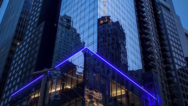 Un edificio de gran altura con fachada de vidrio reflectante y una llamativa franja de luz de neón azul en la base.