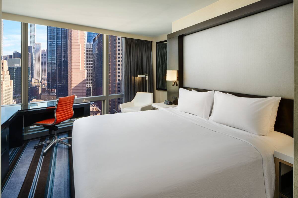 Habitación de hotel con una cama grande, un escritorio con una silla, una lámpara de escritorio y vistas a edificios de la ciudad.