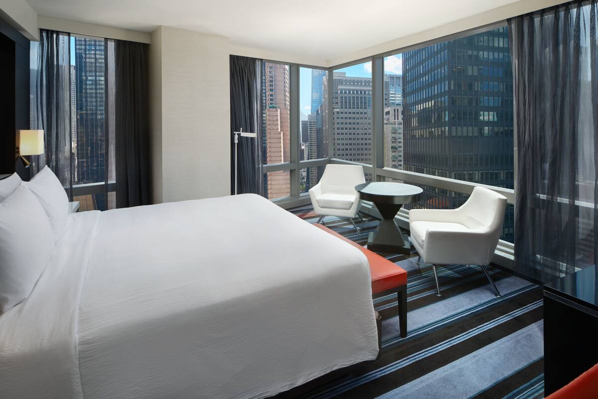 Habitación de hotel con una cama grande, dos sillas blancas, una mesita y vistas a edificios de la ciudad a través de grandes ventanales.