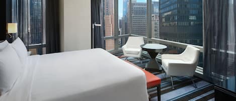 Habitación de hotel con una cama grande, dos sillas blancas, una mesita y vistas a edificios de la ciudad a través de grandes ventanales.