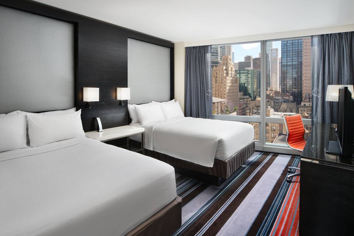 Habitación de hotel con dos camas, un escritorio, una silla y un ventanal con vista a la ciudad.