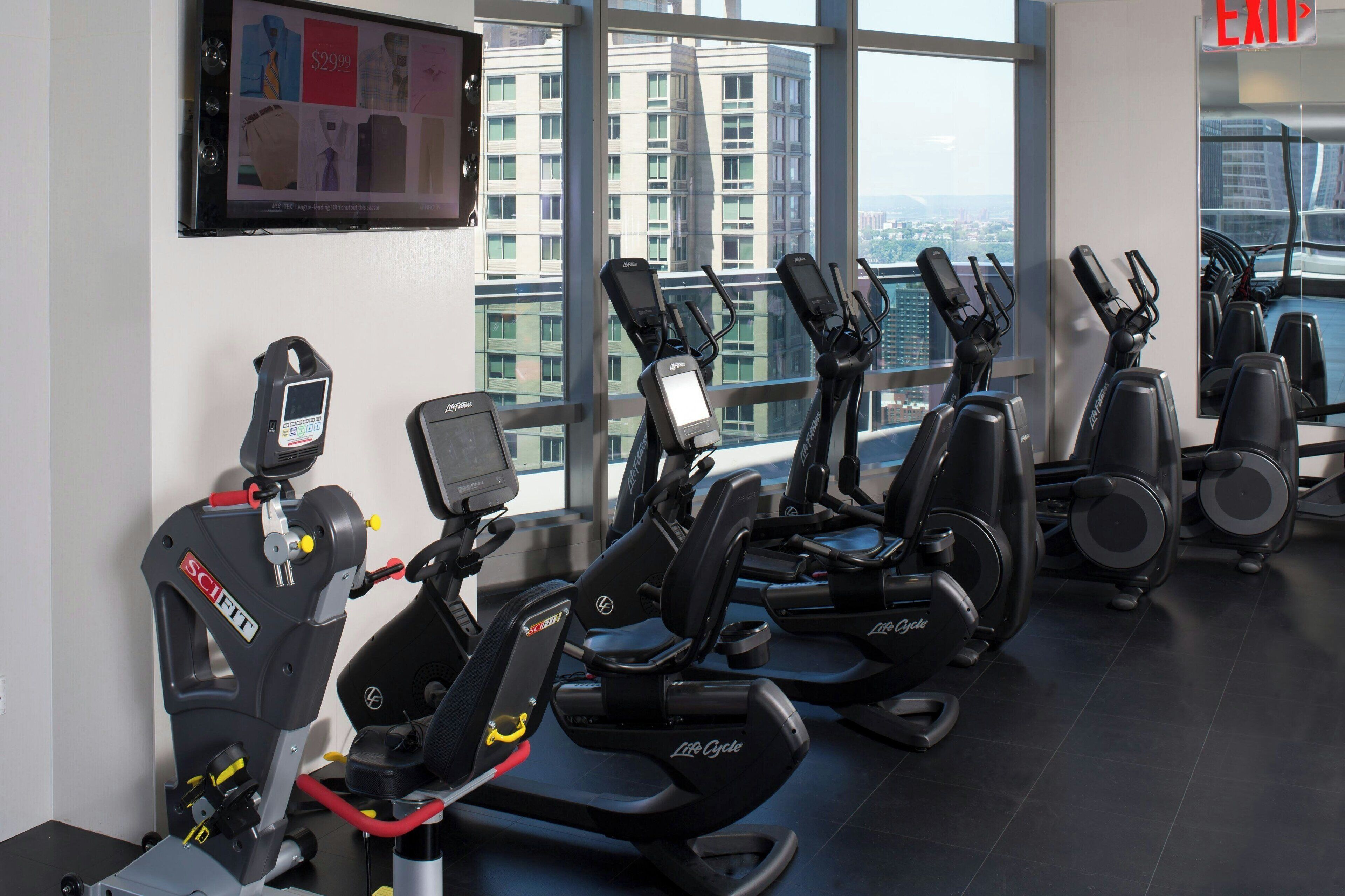Et fitnesscenter med flere løbebånd og andre træningsmaskiner, et digitalt display og et stort vindue med udsigt over byen.