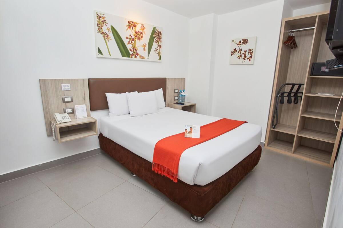 A hotel room with a large bed, a bedside table with a phone and a water bottle, a wooden nightstand, and a closet with shelves.