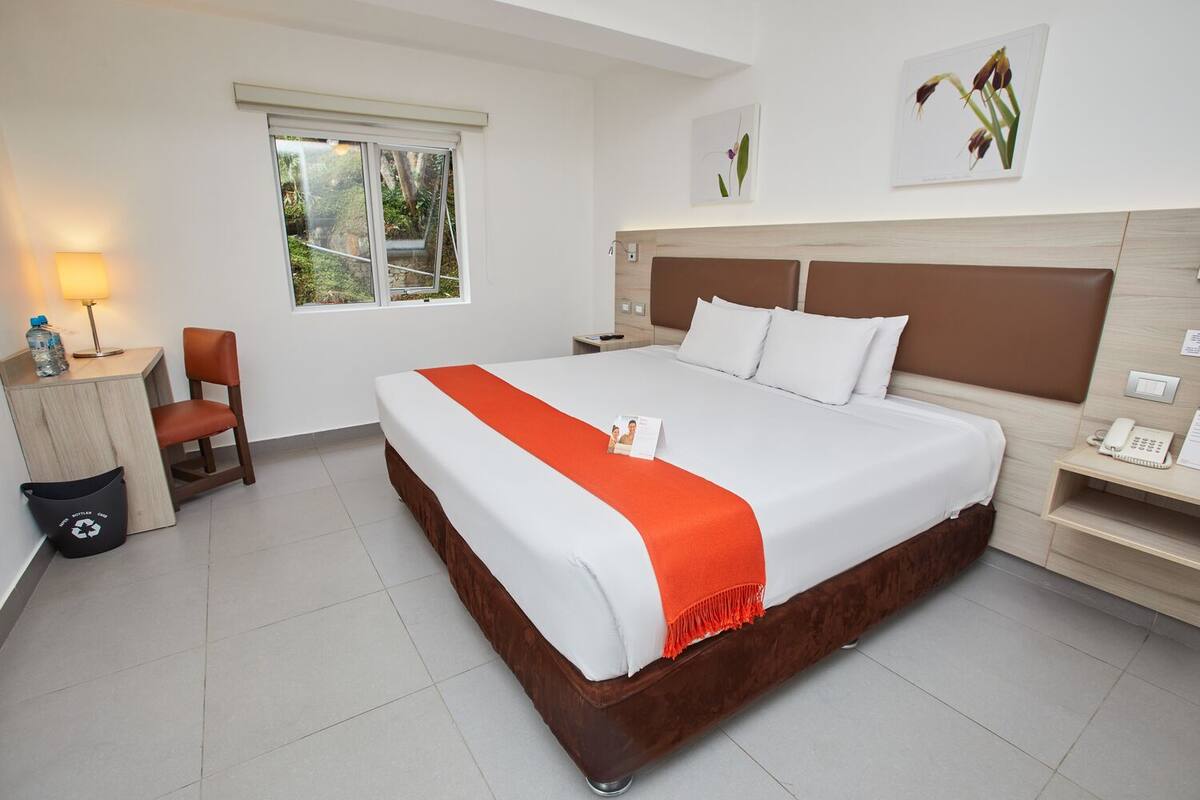 A hotel room with a large bed, a wooden headboard, a desk with a lamp, a chair, and a window with a view of greenery.