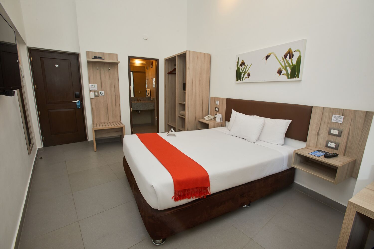A hotel room with a large bed, a wooden headboard, a bedside table with a phone and remote, a small bedside table with a remote, a wooden wardrobe, a wooden bench, and a door.