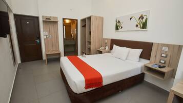 A hotel room with a large bed, a wooden headboard, a bedside table with a phone and remote, a small bedside table with a remote, a wooden wardrobe, a wooden bench, and a door.
