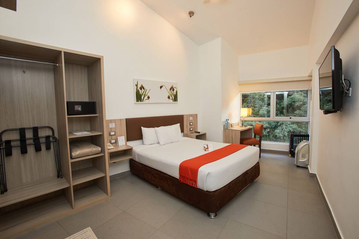 A hotel room with a bed, a desk, a chair, a TV, a window, and a storage unit.