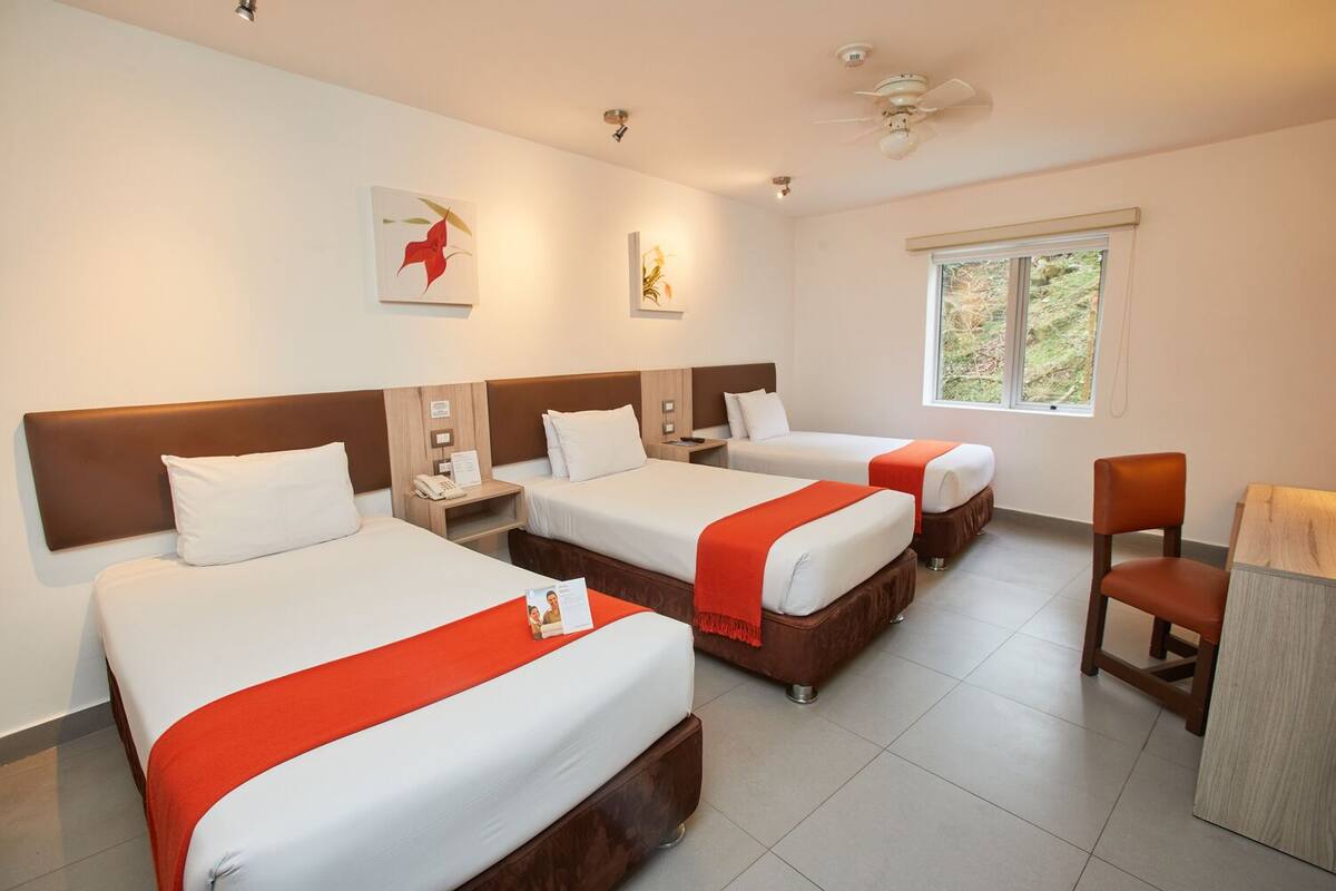 A hotel room with three beds, a desk, a chair, and a window with a view of greenery.