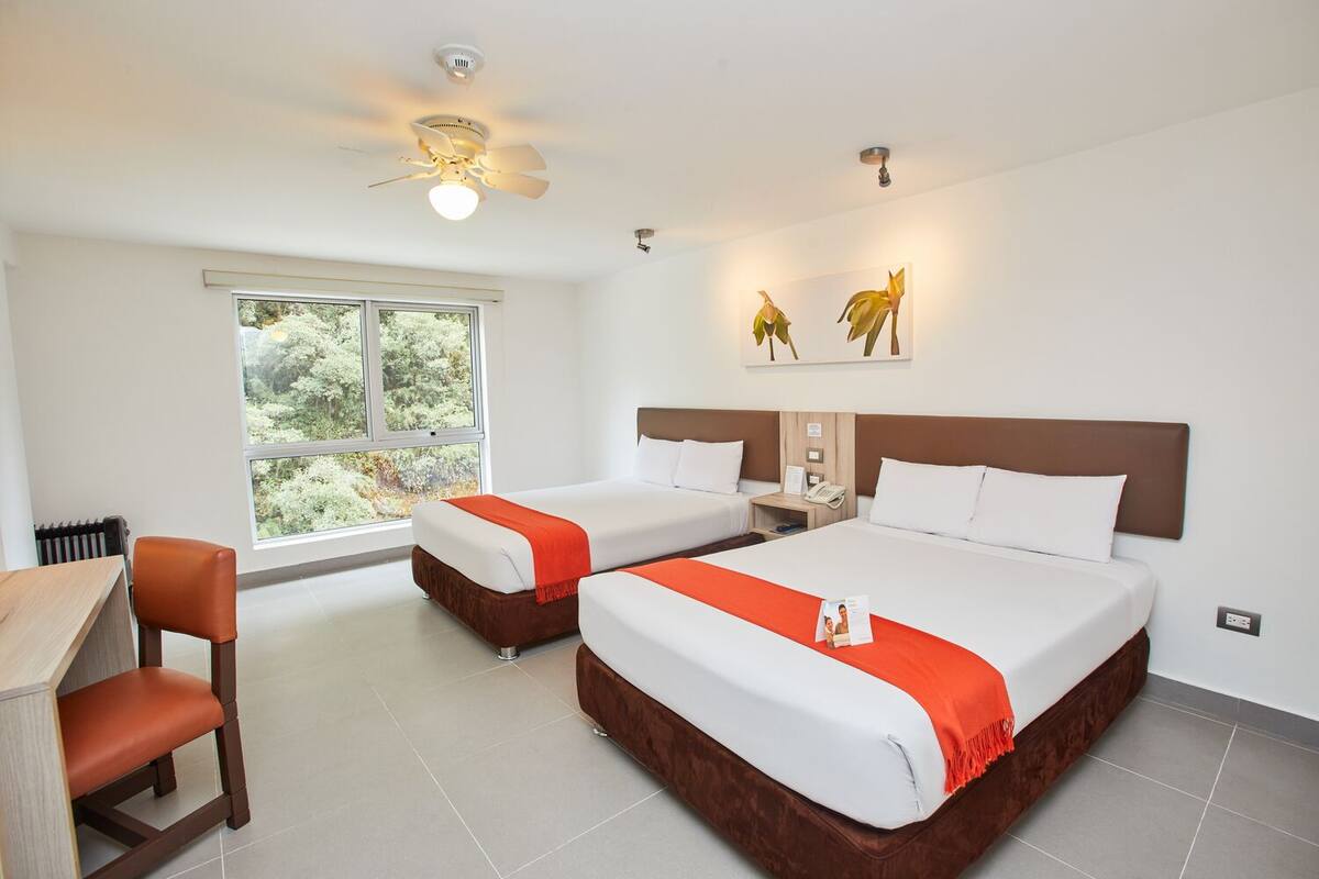 A hotel room with two beds, a desk, and a chair. There is a window with a view of greenery outside.
