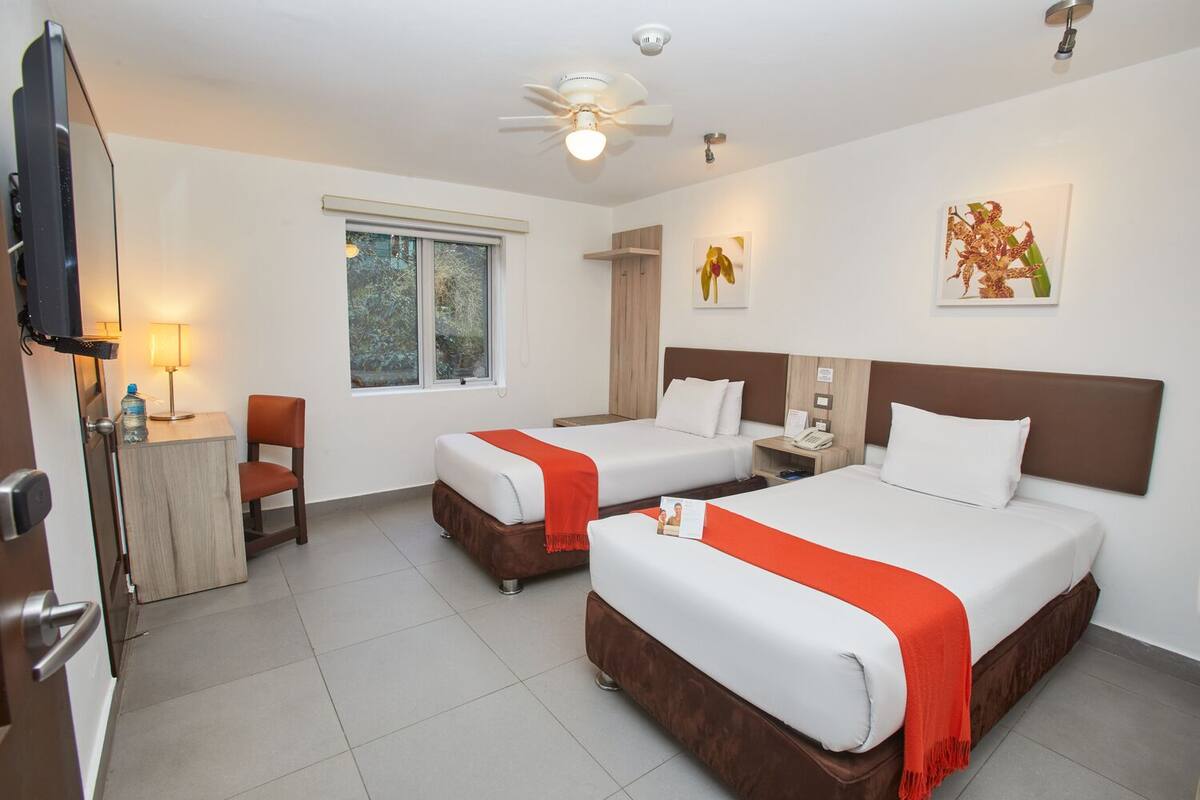 A hotel room with two beds, a desk with a chair, a TV, and a window with a view of trees.