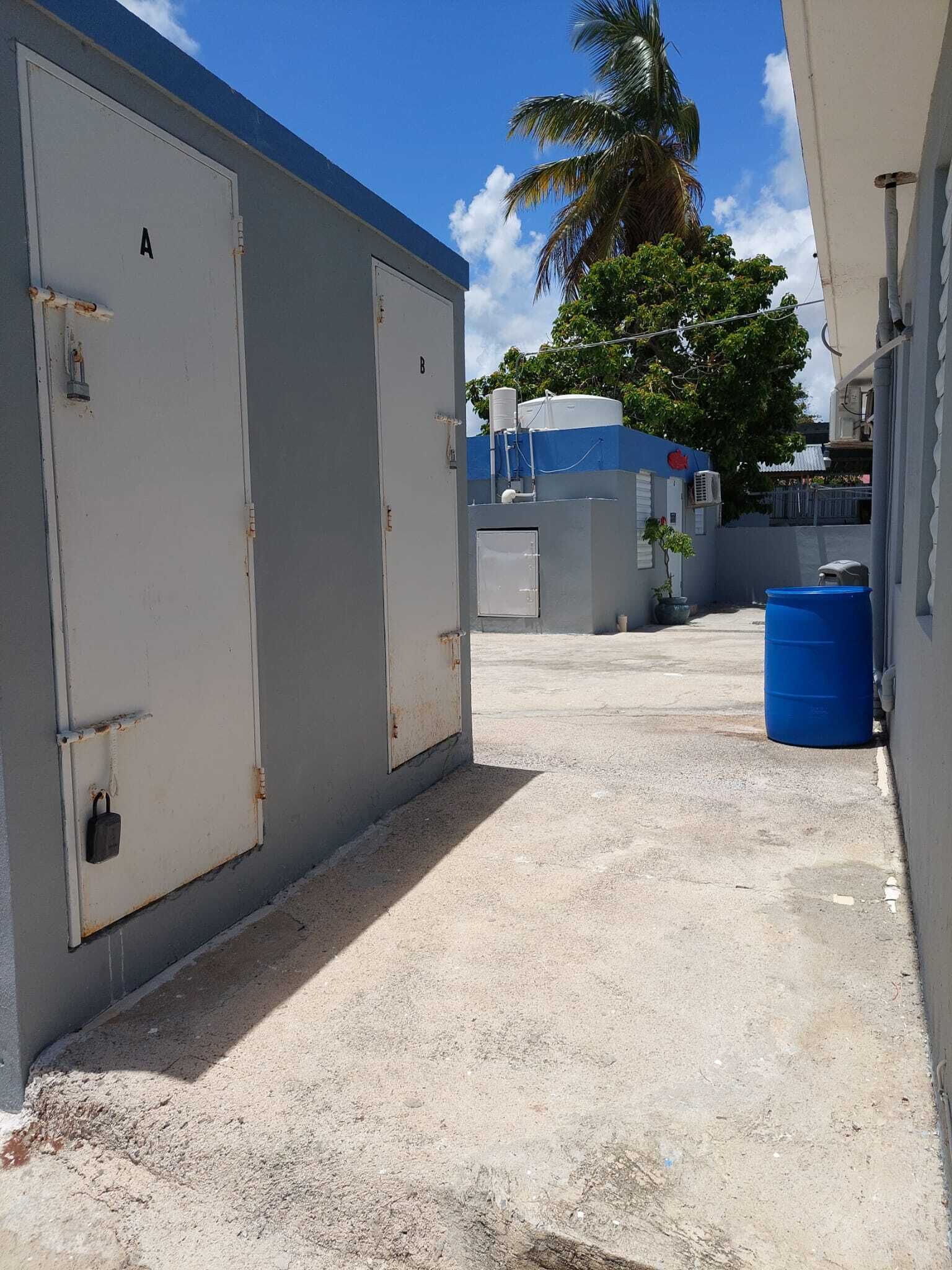 A narrow alleyway with two large, grey utility buildings, a blue barrel, and a palm tree in the background.