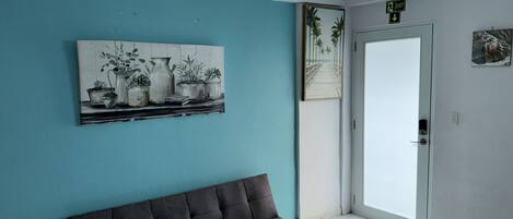 Une pièce avec un mur de couleur turquoise, un canapé gris, une porte avec un panneau indiquant la sortie, et une œuvre d’art encadrée représentant des plantes en pot.